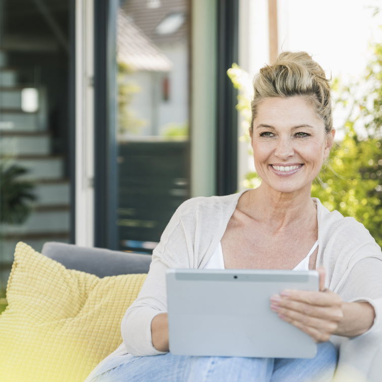 an image of a woman with blonde hair smiling whilst holding an ipad and sat in front of an open door with no blinds 