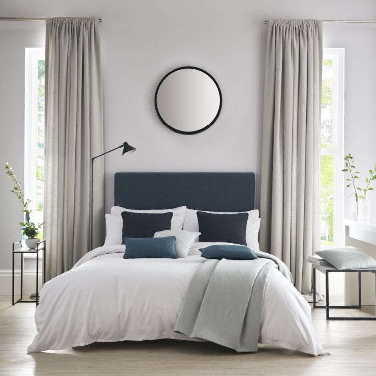 image of white and blue bed next to two windows with cream bedroom curtains 